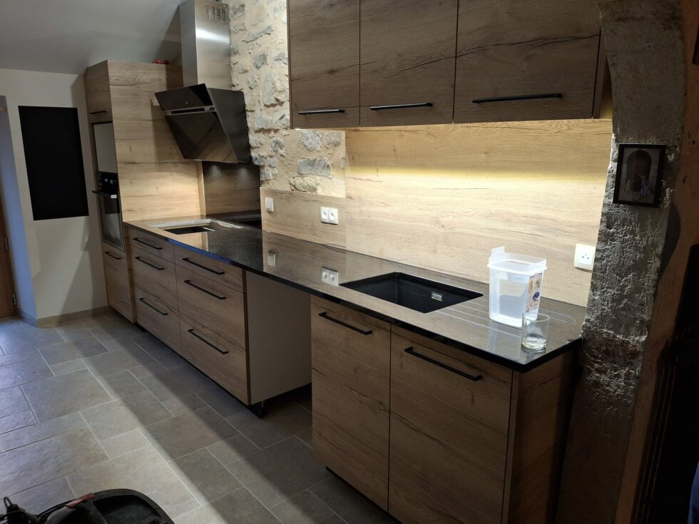 Cuisine linéaire chêne avec plan de travail granit noir contre un mur de pierre